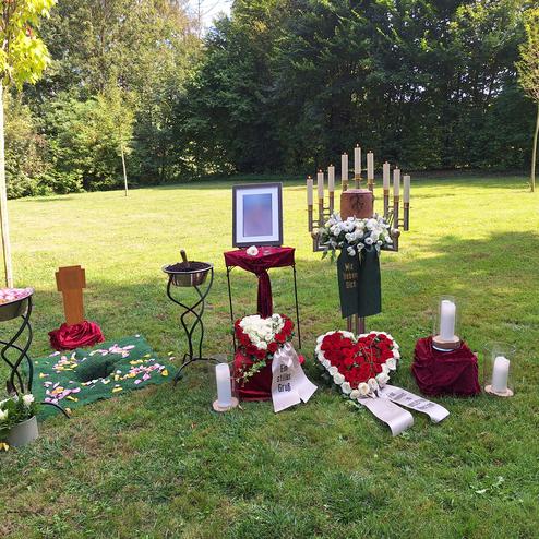 Gedenkst&auml;tte im Freien mit Blumen, Kerzen und einem Bildrahmen auf einem Rasen parzelliert durch B&auml;ume im Hintergrund.
