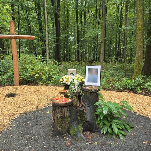 Gedenkst&auml;tte im Wald mit Kreuz, Blumen und Erinnerungsfoto auf Baumstumpf, umgeben von gr&uuml;ner Vegetation.