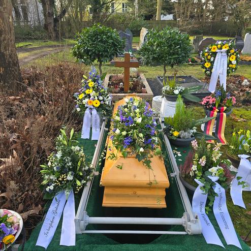 Holzsarg umgeben von bunten Blumenarrangements und Trauerschleifen auf einem Friedhof. Gr&uuml;ne B&auml;ume im Hintergrund.