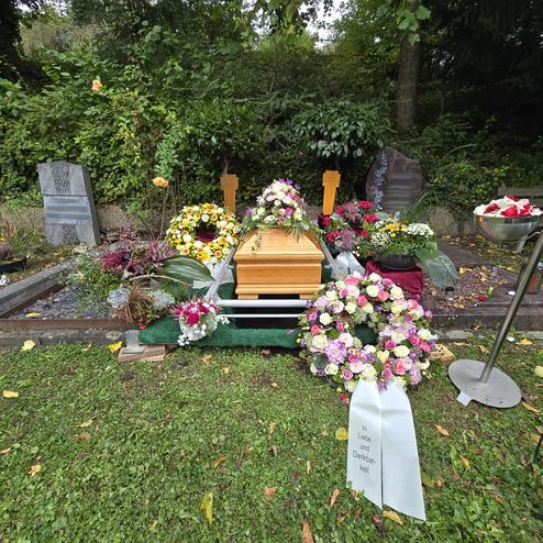 Ein schlicht geschm&uuml;cktes Grab mit einem offenen Holzsarg, umgeben von Blumenkr&auml;nzen und Grabsteinen in einer gr&uuml;nen Umgebung.
