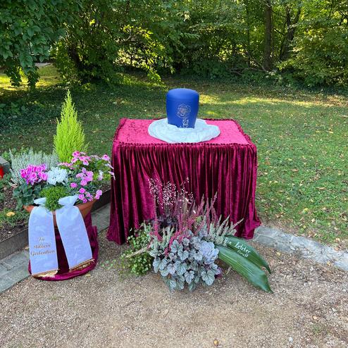 Dekorativer Gedenktisch mit blauer Haube, Blumenarrangements und rotem Tuch in gr&uuml;nem Garten.