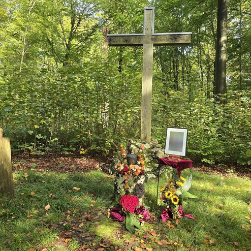 Holzkreuz mit Blumen und Kerze vor einem Bild in einem gr&uuml;nen Waldgebiet. Ein Baumstumpf liegt daneben.