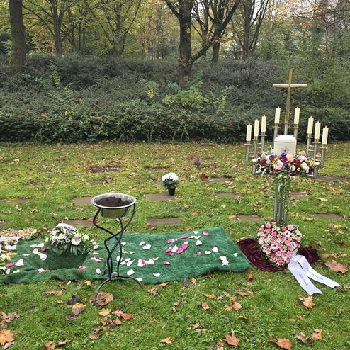 Grabst&auml;tte mit Blumenarrangements, Kerzen und einem Kreuz auf gr&uuml;nem Rasen zwischen herbstlichen B&auml;umen.