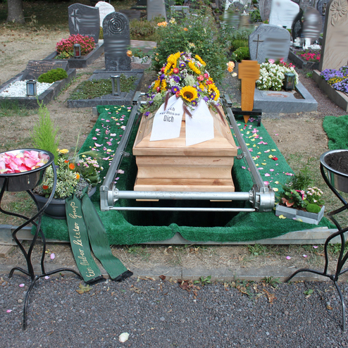 Offenes Grab mit einem h&ouml;lzernen Sarg, umgeben von Blumen, Schalen mit Rosenbl&auml;ttern und Erde auf gr&uuml;nem Teppich.