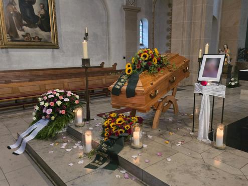 Kiefernholz-Sarg mit Blumenarrangements, Kerzen und einem Bildrahmen in einer Kirche. Florale Dekoration und Trauerschleifen sind sichtbar.