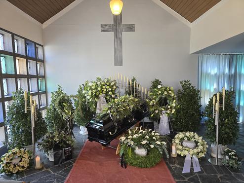 Schwarzer Sarg umgeben von Blumenarrangements, Kerzen und Gr&uuml;npflanzen in einer stimmungsvollen Trauerfeier.