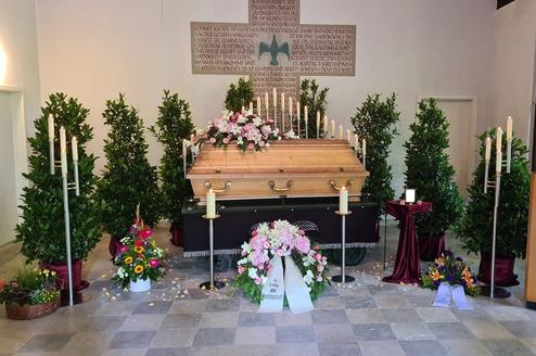 Aufgebahrter Sarg umgeben von Pflanzen und Blumenarrangements in einer Trauerhalle. Kerzen strahlen sanftes Licht aus.