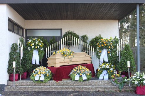 Holzener Sarg auf einer rot belegten Fl&auml;che, umgeben von Blumenarrangements und Kerzen in einer gesch&uuml;tzten Au&szlig;enansicht.