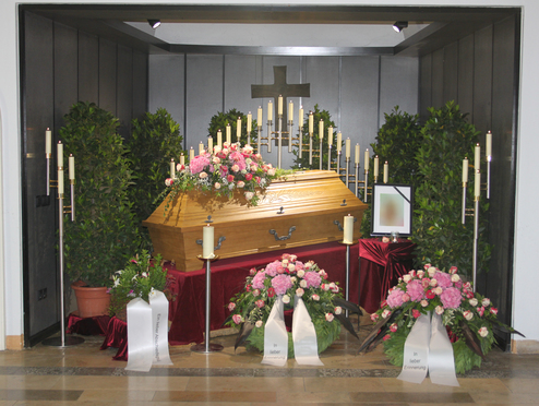 Ein geschm&uuml;ckter Sarg umgeben von Pflanzen, Kerzen und Blumenarrangements in einem Trauerraum.