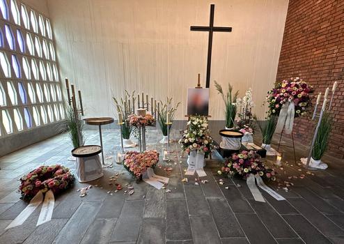 Schmuckvoller Gedenkort mit Blumenarrangements, Kerzen und einem Kreuz in einem lichtdurchfluteten Raum.