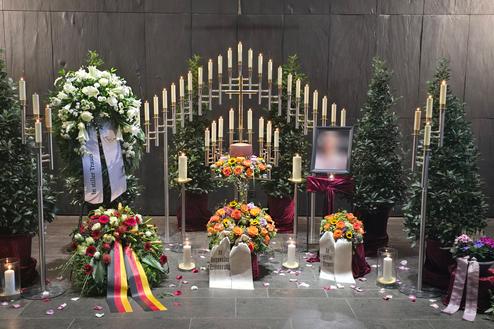 Gedenkst&auml;tte mit Blumenarrangements, Kerzen und einem gerahmten Foto in einer ruhigen Umgebung.