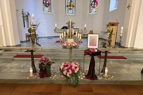 Blumenarrangement und Kerzen auf der Altarstufe einer Kirche mit farbigen Fenstern im Hintergrund.