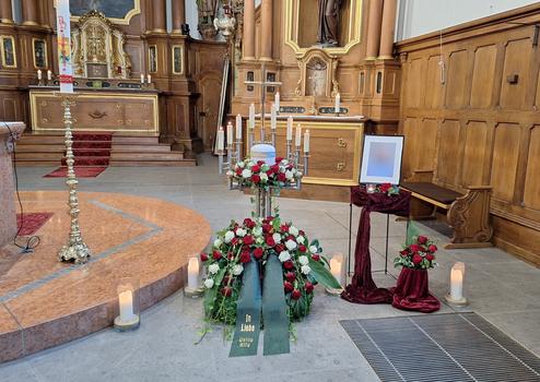 Blumenarrangement mit Rosen und Schleife vor Altar in Kirche, daneben ein Tisch mit Kerzen und einem Foto.