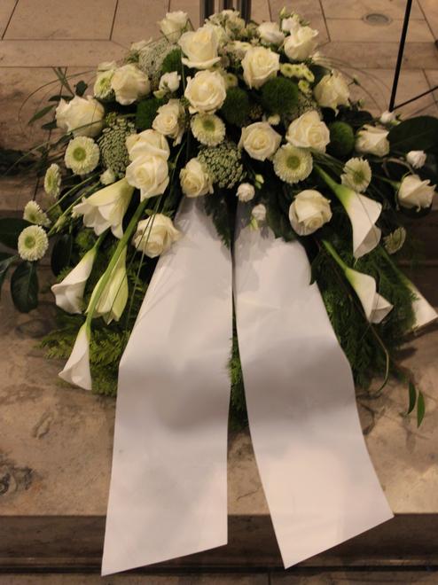 Blumenarrangement mit wei&szlig;en Rosen und Lilien, umgeben von gr&uuml;nem Laub und zwei wei&szlig;en Schleifen.