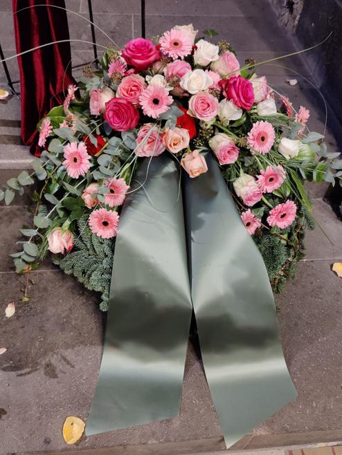 Blumenkranz aus pinken und wei&szlig;en Rosen sowie Gerbera, dekoriert mit gr&uuml;nem Band, auf einem Gehweg platziert.