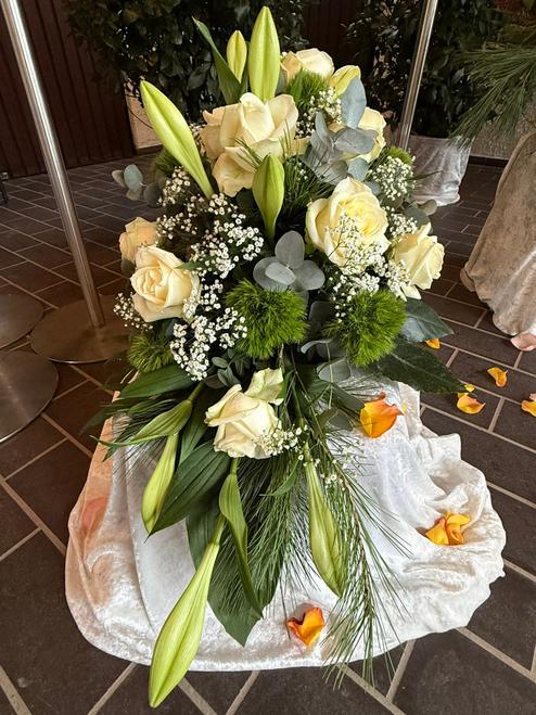 Blumenarrangement mit wei&szlig;en Rosen, Lilien und gr&uuml;nen Akzenten auf einem samtigen Untergrund. Orangefarbene Bl&uuml;tenbl&auml;tter daneben.