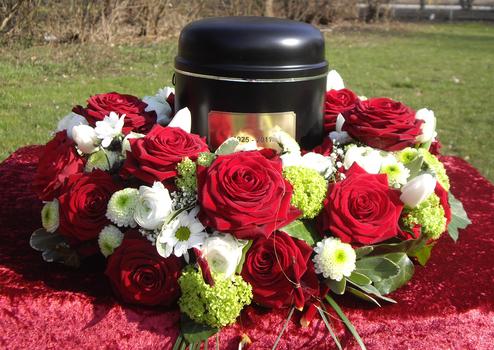 Schwarze Urne umgeben von roten Rosen und wei&szlig;en sowie gr&uuml;nen Bl&uuml;ten auf einem roten Tuch im Freien.