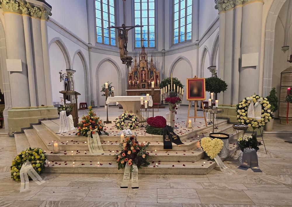 Blumenarrangements und Kerzen auf dem Altar in einer Kirche, hinterlegt mit einem gerahmten Bild.